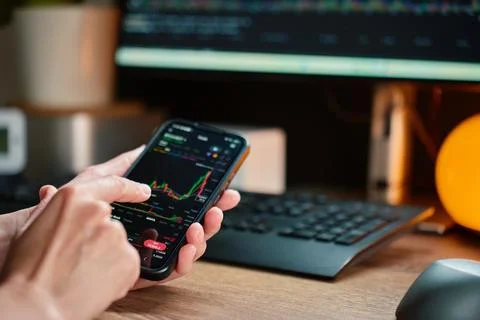 Person using trading app on smartphone at desk with market chart on screen. Stock Photos