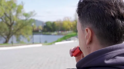 A person using a vape or e-cigarette in the background of an alley in a park Stock Footage 253996015