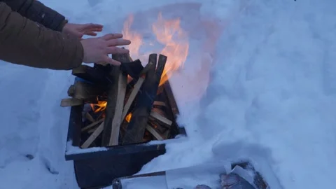 Person warming hands over burning fire in metal smoker box outdoors in deep snow Stock Footage 329579696