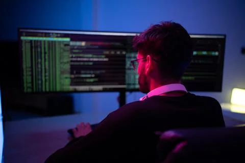 Person working on coding tasks in a dimly lit room with monitors displaying.. Stock Photos