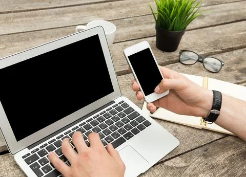 Person working on laptop while using smartphone at wooden desk with plant and Stock Photos