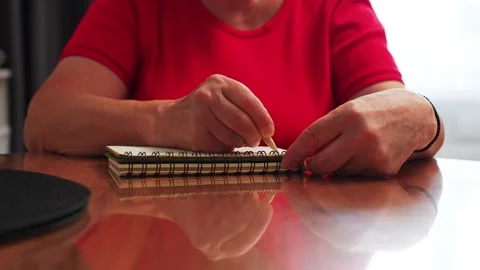 Person writes in a notebook while sitting at a wooden table in a simple room dur Stock Footage 331742736