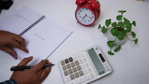 Person writing in a notebook with a calculator, clock, and plant on a desk Stock Footage 317131657