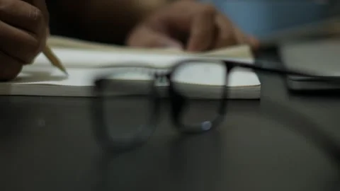 Person Writing in Notebook with Eyeglasses in Foreground Stock Footage 345101648