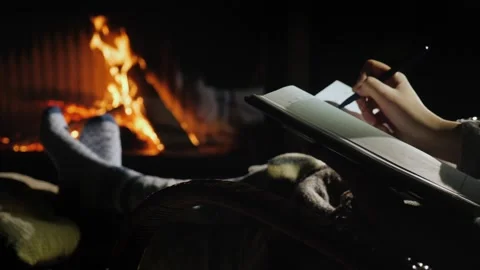 Person writing in a notebook while relaxing by a cozy fireplace at home. Stockbeeldmateriaal 334641597