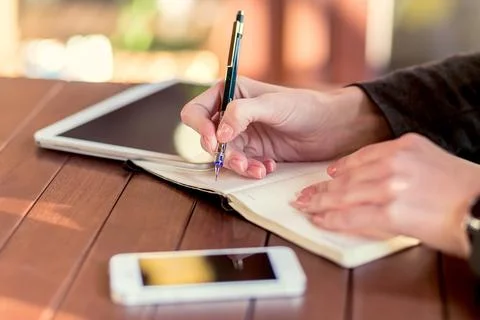 Person writing in a notebook while using a tablet and smartphone on a wooden Stock Photos