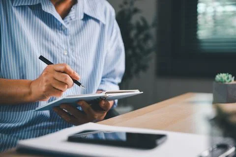 Person writing in a notebook while working beside a smartphone at a modern .. Stock Photos