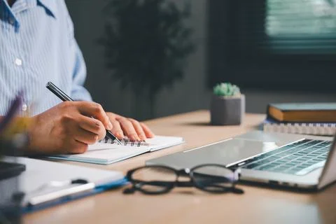Person writing notes in a spiral notebook beside a laptop and eyeglasses at.. Stock Photos