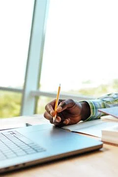 Person writing with pencil while using laptop by the window at a workspace Stockfoto's