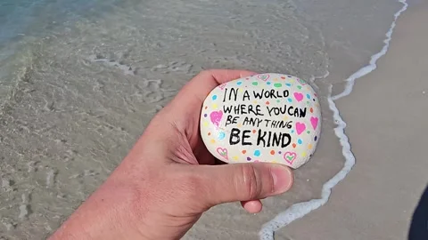 Personal perspective of walker on ocean beach holding be kind kindness rock Stock Footage 328418110