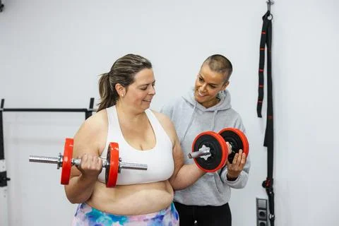 Personal trainer helping client lifting weights in gym for biceps training Stock Photos