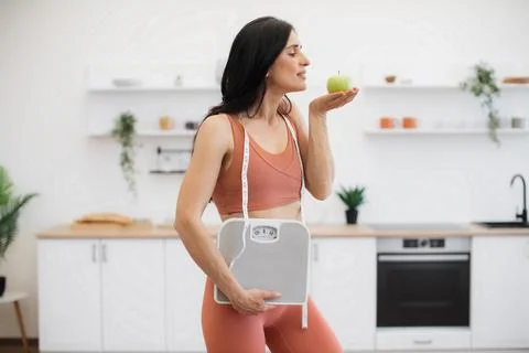 Personal trainer posing with apple and weight scales indoors 写真素材