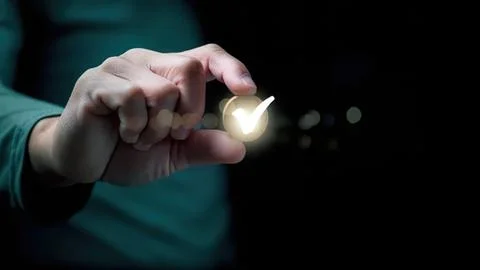 Person's hand holding a glowing checkmark in dark background Stock Photos