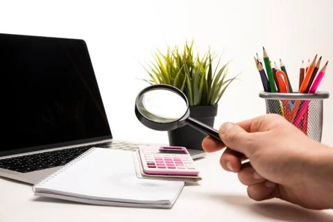 Person's hand holding a magnifier on a desk having a laptop, calculator Stock Photos