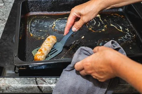 Person's hand lifts a sausage using a spatula from a black baking tray on a k Stock-Fotos