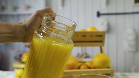 Person's hand pouring pitcher of juice into glass with surrounding oranges - Stock Footage 341262199