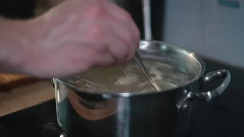 A person's hand stirs a pot of boiling water, adding a spoonful of a thick Stock Footage 338301367