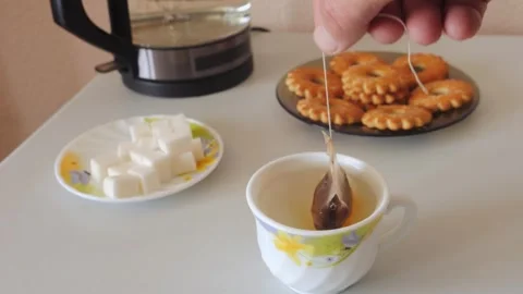 A person's hands dip a tea bag into a cup of hot water on a table with swee.. Vidéo 318415147