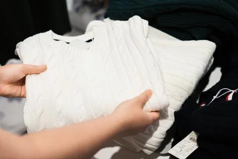 A person's hands hold a folded white cable knit sweater in a clothing store.. Stock Photos