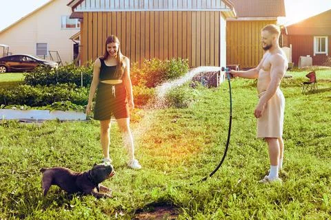 Pet owner waters dog with garden hose, which amuses animal and young woman of Stock Photos
