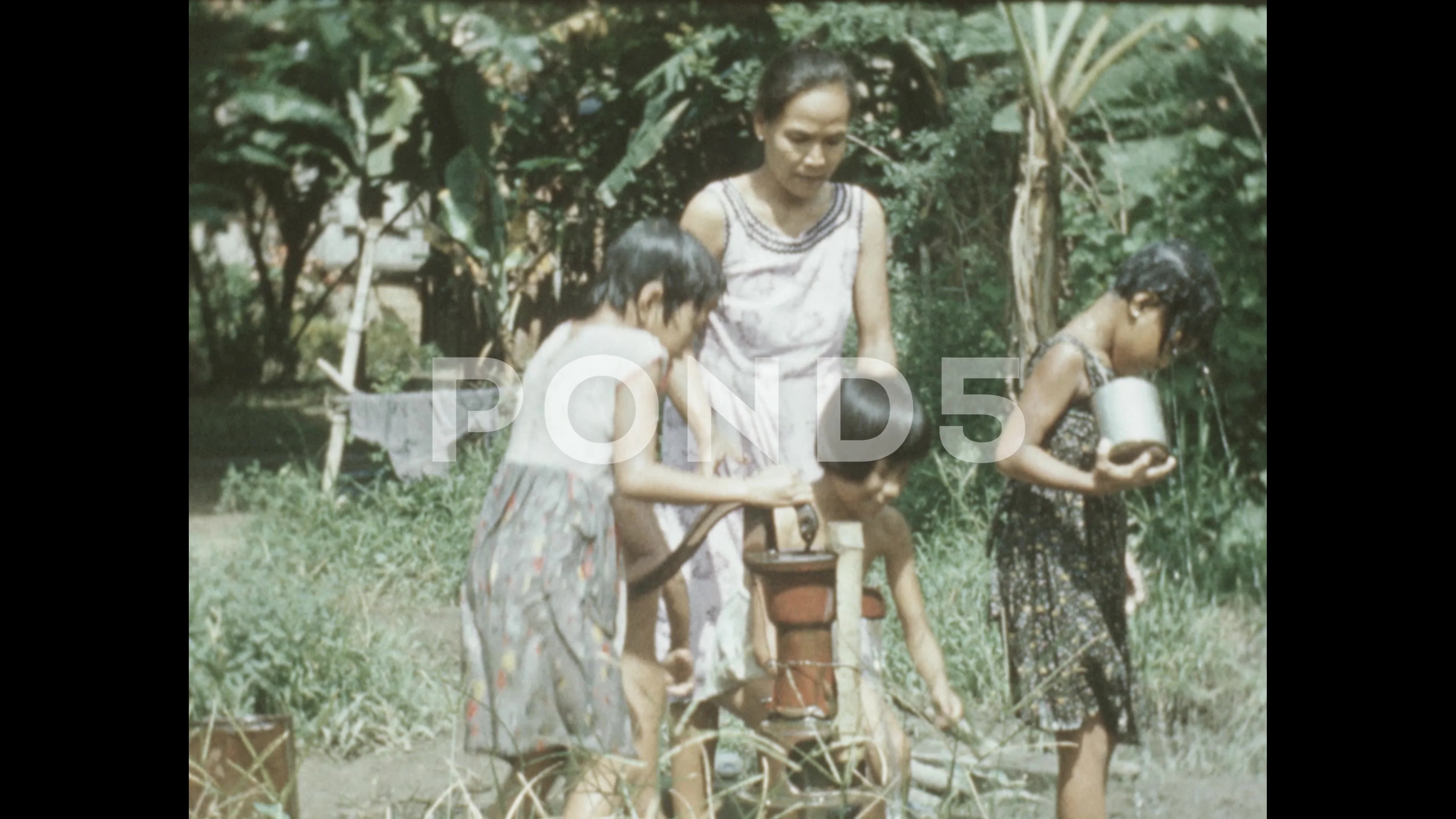 Philippines 1950s: Woman holds a naked boy next to water pump. A girl dumps