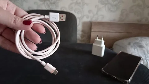 A phone cable in a womans hand, a charging unit and a phone on the table. Phone Stock Footage 319980611