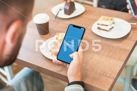 Phone mockup in man hand. Coffee shop table in background with coffee and cake PSD Template