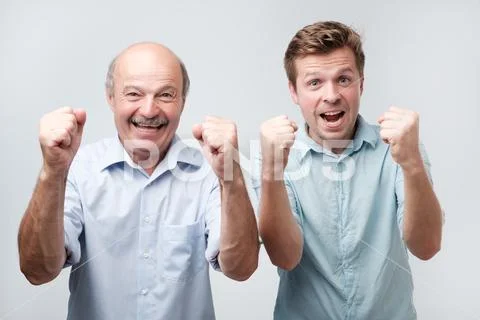 Photo of joyful two young men clench fists and shout with happiness, dressed Stock Photos