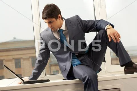 Photo of smart businessman sitting on window-sill with laptop near by and lookin Stock Photos