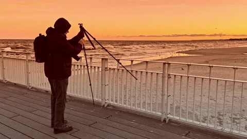 Photographer Setting Up Camera on Pier at Sunset Over Ocean Horizon Видео 297540352