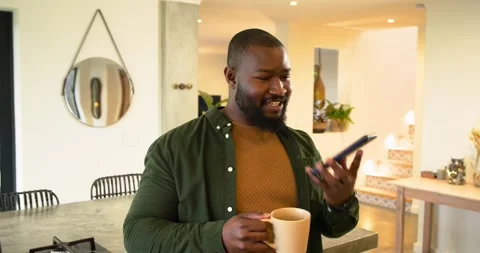 Picking up mug and smartphone in kitchen, African American man laughing and Stock Footage 319779459