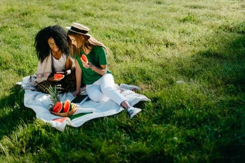 Picnic theme. Two alluring charming african and caucasian girl friends chatting Stock Photos