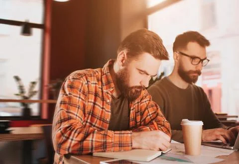 A picture of bearded guy writing down notes in his notebook while his friend is Foto stock
