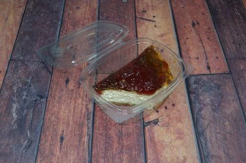 A piece of cheesecake is sitting in a plastic container on a wooden table Stock Photos