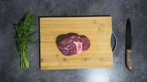Piece of meat on bone falls from above on cutting board on gray kitchen table Stock Footage 299929480