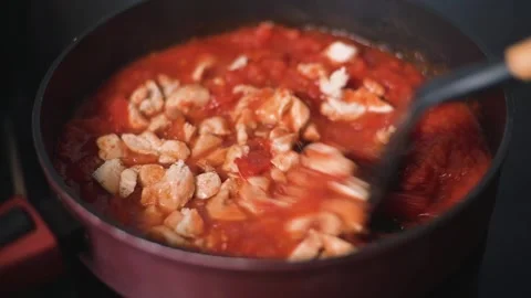 Pieces of chicken are added to a pot of tomato sauce on a stove in a kitchen. 動画素材 344667488