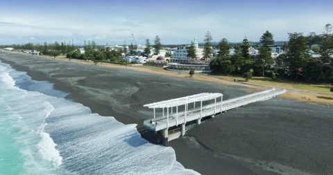 A pier extends over a black sand beach in Marine Parade, Napier, New Zealand Stock-Footage 332816678