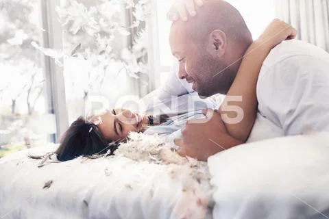 Pillow feathers falling around playful, affectionate couple on bed Stock Photos