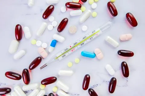 Pills of different forms and colors and medical thermometer on white marble b Stock Photos