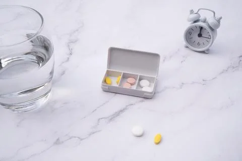 Pills inside a pillbox on a table, next to a clock and  a glass of water. Stock Photos
