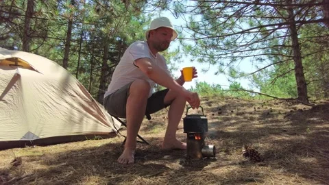 Pine Forest Camp: Man Sipping Tea, Kettle on Wood Burner Near Tent Stock Footage 299261125