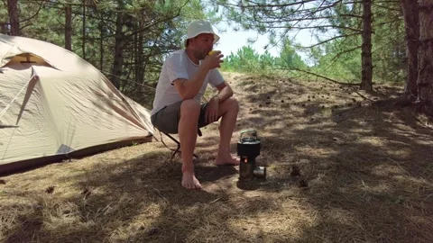 Pine Forest Camp: Man Sipping Tea, Kettle on Wood Burner Near Tent Stock Footage 303505788