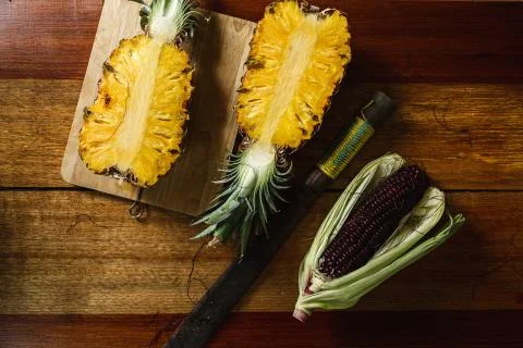 Pineapple and purple corn lying on a wooden table next to a large knife Stock Photos
