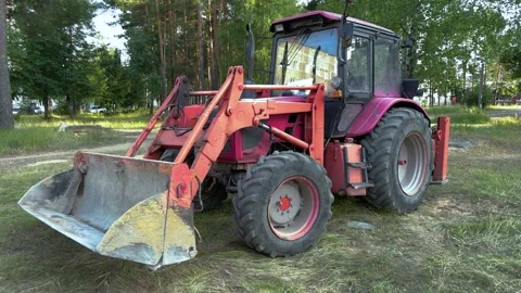 Pink and red tractor with front loader bucket parked on grass near forest Видео 338201484