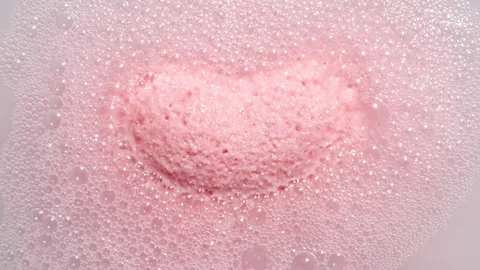 Pink bath bomb ball in the shape of a heart dissolves in water with white foam Stock Footage 253774615