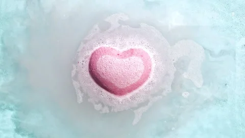 Pink bath bomb ball in the shape of a heart dissolves in water with white foam Stock Footage 255676195