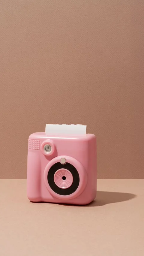 A pink camera rests on a textured brown backdrop as paper emerges in stop motion Stock Footage 330376390