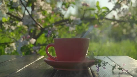 Pink ceramic cup with steam and flowers on wooden table, spoon stirring inside, Stock Footage 334757572