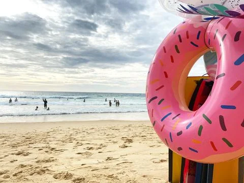 Pink donut-shaped inflatable float placed on sandy beach with people swimming in Stock Photos