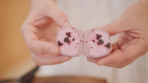 Pink mochi split revealing berry filling held by white hands over wooden board Stock-Footage 347328949